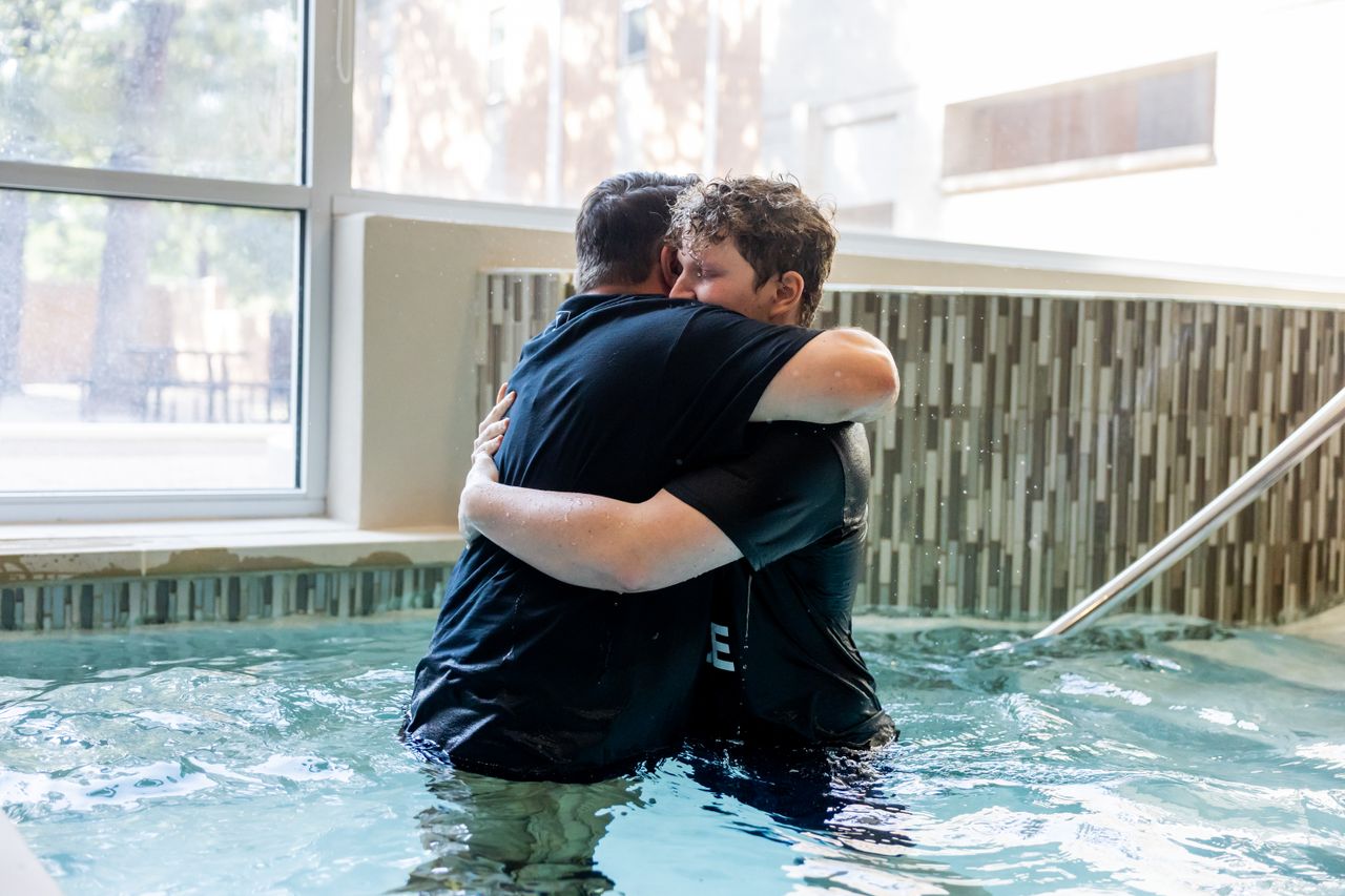 ours-journey Two men embrace after baptismal ceremony in an indoor pool, symbolizing faith and community.
